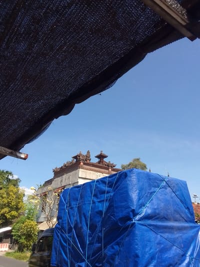 The image depicts an outdoor scene in Gianyar, Indonesia, under a clear blue sky. The top third of the frame is occupied by a dark, coarse mesh material, likely a shade net or canopy, with a visible weave pattern. A metallic fixture extends from beneath this netting on the upper left.
In the midground, a traditional Balinese building or temple structure is prominent on the left, featuring multi-tiered, ornate dark brown roofs. To its right, partially obscured by a blue tarp, is a structure with exposed concrete and rebar, suggesting construction or renovation.
The lower half and right side of the image are dominated by a dark-colored vehicle, likely a pickup truck or small lorry, carrying a substantial load. This load is completely covered by a taut, bright blue tarp, which is secured by a network of blue ropes crisscrossing its surface. On the far left, a section of a paved road is visible, bordered by green grass and lush vegetation, including trees with green leaves and some bearing yellow flowers. A white and orange striped traffic barrier is partially visible near the road's edge. No individuals are discernible in the scene.