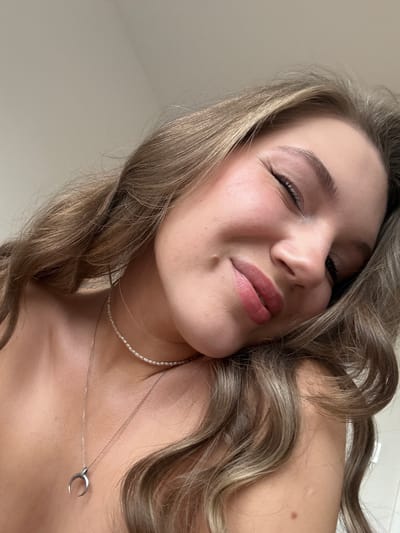 A low-angle close-up captures a woman with shoulder-length, wavy brown hair. Her face is turned slightly to the side, with one eye closed in a wink. She wears makeup including blush and lipstick. Around her neck is a pearl necklace and a silver chain with a crescent moon pendant. A mole is visible on her left cheek and upper chest. The background is a plain, light-colored surface. The image does not contain any information about the location being Vozdovac Urban Municipality, Serbia.