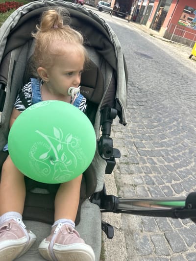 This is a vertical, eye-level shot of a toddler lying in a stroller on a cobblestone street. The toddler has blonde hair pulled up in a bun and is wearing a denim overall dress over a black and white striped shirt, white socks, and pink high-top sneakers. A pacifier is in the toddler's mouth. A large, light green balloon with a white floral design is resting on the toddler's chest.
The background shows a cobblestone street with buildings on the left side of the image. The buildings appear to be shops or restaurants, with a sign that reads "25 Мебел" in Cyrillic script, which translates to "25 Furniture" and is likely an advertisement for a furniture store. The scene suggests an outdoor setting in a town or city. The lighting indicates it is daytime, possibly with overcast skies, as there are no strong shadows. The overall mood seems peaceful, with the toddler appearing calm and content in the stroller.