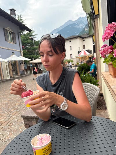 A woman is seated at a table, holding a cup of ice cream and a spoon, in what appears to be an outdoor cafe or restaurant setting in the city of Molveno, Italy. She is wearing a dark gray t-shirt and a watch on her left wrist. A second cup of ice cream is on the table in front of her. In the background, buildings, trees, and mountains are visible, along with a carousel. The pavement is made of cobblestones.