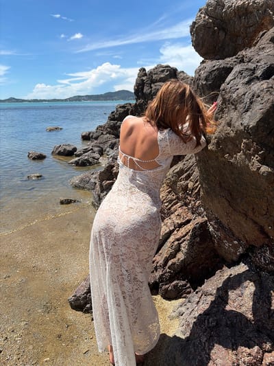 A woman with long, reddish-brown hair, viewed from the back, leans against dark, jagged rocks near the ocean. She is wearing a white lace dress with an open back, adorned with a delicate pearl-like embellishment. The dress is long and form-fitting, with intricate floral lace patterns visible through the fabric. Her right hand is resting on the rocks, and her left arm is bent. The scene appears to be set on a rocky shore with some sand. The ocean is calm and blue, reflecting the bright sky, which is dotted with white, wispy clouds. In the distance, a faint outline of mountains or hills can be seen. The lighting suggests it is daytime, likely sunny with some shadows cast by the rocks. The overall mood is serene and peaceful, with a focus on the natural beauty of the location and the woman's pose. There is no visible text in the image. The location context is Baan Bo Phut, Thailand.