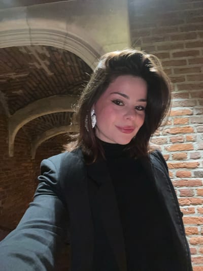 A woman with brown hair and a black turtleneck smiles into the camera. She is wearing silver earrings. A brick wall and arched brick feature is visible in the background. The image is taken from a slightly downward angle. Her left arm is visible in the lower right corner.