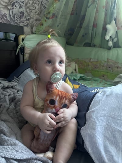 A young child with blond hair pulled into a small ponytail and blue eyes is sitting on a bed, clutching a stuffed cat toy. The child has a light blue pacifier in their mouth. The child is wearing a yellow tank top. The bed has dark blue and gray patterned bedding. In the background, there are sheer green curtains with a pattern of animals, and a white stuffed star hanging. A floral wallpaper is visible on the wall behind the bed. The overall environment appears to be a child's bedroom. The location is identified as Sayanogorsk, Russia.