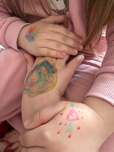The image shows a close-up of a child's hands with colorful drawings and what appears to be a temporary tattoo on the back of the left hand. The child is wearing a pink long-sleeved shirt. The drawings on the right hand include a heart shape and scattered red dots, with a light blue smudge. The temporary tattoo on the left hand is a multi-colored flower. The background is out of focus and appears to be part of the child's clothing. The image does not provide any information or context that would allow for its location to be identified as Valozhyn, Belarus.