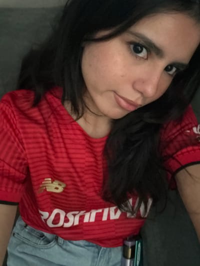 A person with dark hair is lying down and looking at the camera. The person is wearing a red soccer jersey with "POSITIONS" written in white. The person is also wearing light blue denim jeans. There is a gray surface below the person, and an object that appears to be a tube of some kind is visible next to the jeans. The location cannot be verified as College Station, United States of America from this image.