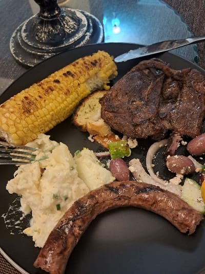 A black circular plate displays a cooked meal, featuring a grilled lamb chop, a charred corn on the cob, a portion of potato salad with green flecks, a curved grilled boerewors sausage, and a side salad containing red onion, dark olives, and crumbled feta cheese. A silver fork is partially visible in the potato salad, and a silver knife rests on the plate's upper edge. In the background, a distressed black lamp base is visible on the upper left, and a blurred blue light source appears on the upper right. The plate rests on a dark, textured surface. This scene is located in Mitchells Plain, South Africa.