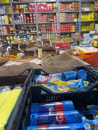 The image displays the interior of a densely stocked retail establishment, likely a grocery or spice shop, within Ksar-el-Kebir, Morocco. The scene is dominated by shelves packed with a diverse array of products, bulk commodity displays, and foreground crates containing household goods.

In the background, multiple grey metal shelves extend from floor to ceiling, filled with packaged food items. Products include numerous red-labeled Nescafe coffee jars, various canned goods, jars of preserves and pickles, boxed items, and stacked red and white cigarette packs.

The midground features a market-style display with large, open wooden or plastic bins containing bulk goods. Prominent mounds of various light brown and grey-green spices or seeds (e.g., cumin, fennel) are central. Other bins hold dried fruits, such as dark dates and prunes, along with piles of almonds and other nuts. A small Nutella jar, a light-colored cylindrical container, and a light blue scoop are visible among these bulk items. A black weighing scale arm protrudes from the left side. On the right, a vibrant pile of red powdered spice is displayed.

In the immediate foreground, two black plastic crates are partially visible. The left crate contains multiple clear-plastic-wrapped yellow sponges, each featuring a green abrasive layer. The right crate holds several rolls of blue plastic trash bags, labeled "Pure Bag" and "TRASH BAGS MEDIUM 35L," alongside individually packaged circular plastic items (yellow, pink) and coils of blue string or rope. No individuals are present in the scene.
