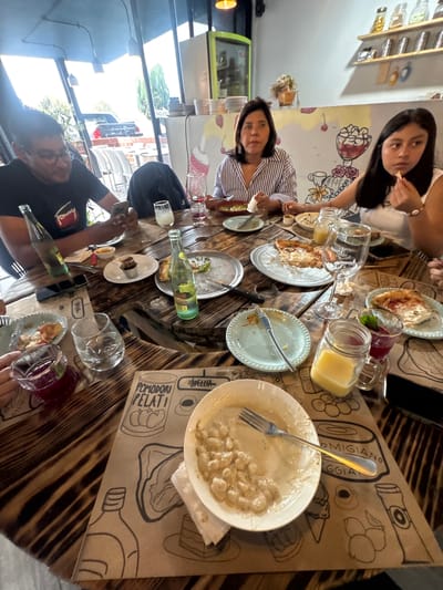 A group of people are seated around a wooden table at a restaurant in San Andrés Cholula, Mexico. The table is covered with brown paper featuring food-related illustrations, and there are several dishes on the table, including pasta and pizza, along with various beverages in glasses and bottles. Several people are interacting and eating. Some people are holding forks and knives. The restaurant interior is visible in the background, and the exterior environment can be partially seen through the windows.