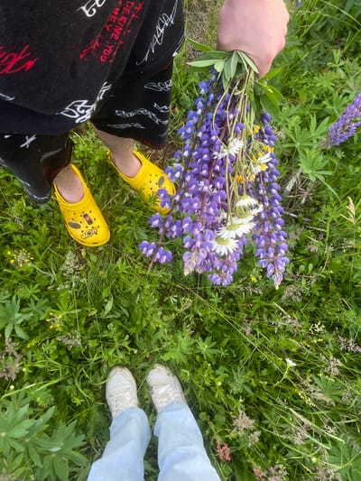 In the city of Kuyeda, Russia, a close-up, overhead shot captures a person's legs and feet standing amidst a field of lush green grass. The individual on the left wears black shorts with a white and red graphic print and bright yellow Crocs. The person on the right is clad in light blue jeans and white sneakers. A hand holds a bouquet of vibrant purple lupine flowers, interspersed with white daisies and yellow wildflowers. The overall scene suggests a serene outdoor activity, likely a walk or gathering of flowers in a natural setting.