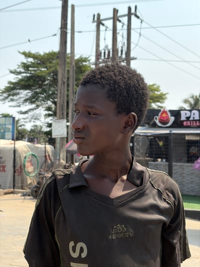 The image displays a young male subject in Ikota, Nigeria, shown in a close-up, three-quarter profile, facing towards the left side of the frame. The subject has dark skin, short dark curly hair, and wears a dark, possibly black or dark brown, collared shirt with a V-neck, featuring "EBS SPORTS" printed in a lighter color on the chest. His expression is neutral. The scene is an outdoor, sunlit urban environment. In the background to the left, a light-colored tarp-covered structure, a blue rectangular sign, a green hoop-like object, and part of a cart are visible. To the right in the background, a commercial building with a black facade features white lettering "PA..." and "GRILL" along with a logo depicting a flaming cup. This building is behind a metal fence. Multiple wooden utility poles with overhead wires and insulators are prominent throughout the background, along with green trees and foliage. The ground in the foreground appears to be an unpaved or dusty surface. No direct interactions are depicted.