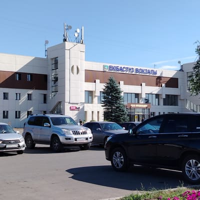 The image shows the Ekibastuz railway station in Ekibastuz, Kazakhstan. A modern, white and brown building with blue lettering that reads "ЕКІБАСТУЗ ВОКЗАЛЫ" (Ekibastuz Station) is visible. The building has large windows and several antenna towers on its roof. In front of the building, there is a parking lot with several cars, including a silver SUV, a dark SUV, and a silver sedan. Some greenery and flowers are also present in the foreground. The sky is clear and blue, indicating a sunny day.