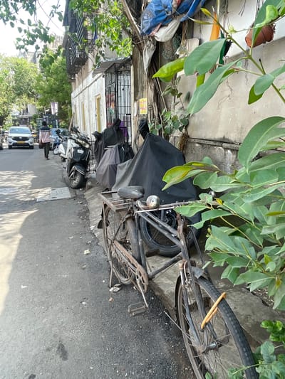 A street scene in Mumbai, India, features a paved road with parked scooters and a bicycle. The buildings on the right are weathered and appear to be residential or commercial. Lush green foliage, including a large tree with broad leaves, overhangs the sidewalk and a portion of the street, casting shadows. Several scooters are parked along the sidewalk, some covered with dark tarps. A vintage bicycle with a metal basket and a rusty frame is prominently featured in the foreground, secured with a yellow lock. A white car is visible in the background, driving away from the viewer. A person in a pink and blue shirt walks away from the car on the left side of the road. The overall impression is a busy urban environment with a blend of nature and everyday life.