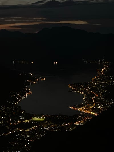 The image presents an elevated, wide-angle nocturnal view of Čavori, Montenegro. A vast, dark body of water, likely a bay or lake, occupies the central and lower portions of the frame. The irregular coastline is delineated by numerous warm-toned artificial lights, forming an illuminated crescent that outlines the urban or semi-urban area. These lights vary in intensity and density, with some clusters suggesting more developed zones or larger structures, including a distinctly brighter, somewhat rectangular area of lights in the bottom center, possibly a sports field. The lights reflect faintly on the calm surface of the water, creating subtle streaks and points of illumination. In the distant background, a dark, undulating range of mountains is silhouetted against the low horizon. Above the mountains, the sky transitions from a faint, dark orange or brown glow along the horizon to a deeper, almost black expanse overhead, with dark, irregular cloud formations visible. No discernable individuals, specific vehicles, or direct actions are visible within the frame due to the distance and darkness.