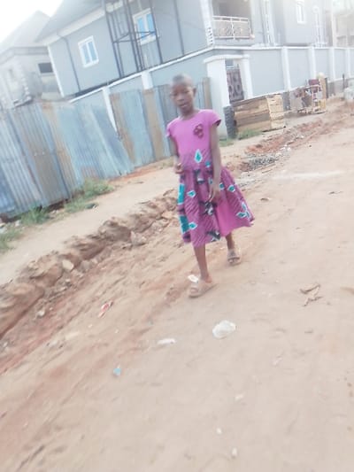 A young girl walks on an unpaved, reddish-brown dirt path in Owerri, Nigeria, angled slightly towards the viewer. She is wearing a short-sleeved, pink dress featuring a lower section with a pattern of teal and white geometric shapes on a darker pink or maroon background, and light-colored sandals. A small, dark embellishment is visible on the upper left chest of her dress. To her left, a shallow earthen trench runs parallel to the path. In the background, a weathered corrugated metal fence stands to the left. Behind her and to the right, a multi-story residential building, painted light grey, is visible with a balcony and an external dark metal structure. Stacked wooden pallets and other debris are located near the building's base. Scattered refuse is present on the dirt path. The scene is illuminated by natural daylight.