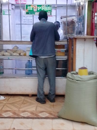 The image shows a man standing in front of a counter inside a small shop in Kivaywa, Kenya. The man is wearing a blue jacket and gray pants. He is looking down at a counter that has a large green bag sitting on the floor in front of it. Behind the counter are shelves with various objects including a bottle of water. The counter is made of wood and has a white painted top. The floor is tiled with brown and beige tiles. The shop's walls are white and have a sign that reads "M-PESA." The location based on the coordinates is a small shop on a street in Kivaywa, Kenya.  The image is taken from a slightly low angle, giving the viewer a perspective from the man's point of view.  The photo is in focus and the lighting is dim. The image was taken on a sunny day.