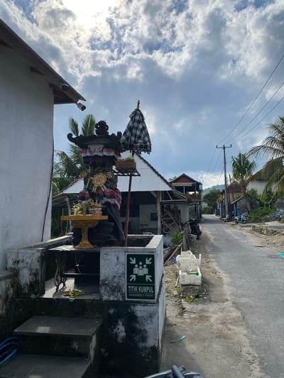 This media file captures a scene in Nusa Penida, Indonesia. The primary subject is a traditional Balinese statue, adorned with a black and white checkered umbrella, sarongs in red and white, and decorative offerings. The statue is positioned in front of a building with a corrugated metal roof and appears to be part of a local shrine or decoration.
Behind the statue, a paved road runs through a village. The setting includes residential structures, some appearing under construction or in disrepair, with visible wooden beams and scaffolding. Tropical vegetation, including palm trees, is present throughout the scene, indicating a warm climate. Electrical wires are strung across utility poles, a common sight in many Indonesian villages.
To the right of the statue, a concrete structure displays a green and white sign with text that reads "TITIK KUMPUL ASSEMBLY POINT". The sign also features an icon with arrows pointing towards a group of people, suggesting a designated meeting or evacuation point. Nearby, there are white plastic containers and some miscellaneous debris, hinting at ongoing activity or a less-than-pristine environment. A scooter is parked further down the road.
The sky is partly cloudy, with patches of bright sun breaking through, suggesting it is daytime, likely late morning or early afternoon. The overall atmosphere appears calm and possibly humid, typical of a tropical locale. No people are directly visible interacting with the statue or the immediate surroundings, lending a sense of quietude to the scene. The composition is a vertical orientation.