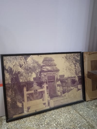 The image shows a framed black and white photograph of a stone building with a wooden door. The building appears to be a temple or shrine, with a tiered roof and stone walls. The photograph is displayed on a wall in a room with a tiled floor. The wall is white with a light grey tint, and the tiled floor is grey with white speckles. There is a second framed picture to the right of the first. The first picture is mounted in a black frame, while the second is mounted in a wooden frame with a light brown hue. The second picture has a white background and an indeterminate subject matter. There are no people or objects in the room besides the two framed pictures, which are against the wall. The image is taken from a perspective looking directly at the pictures. The photograph was likely taken in Surakarta, Indonesia, at the geographic coordinates of -7.559 latitude and 110.783 longitude.