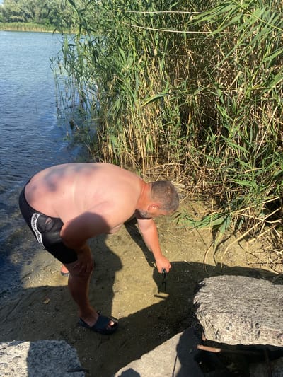 A man, appearing to be in his late 40s or 50s, with a visible abdomen and back, wearing black swim trunks with white lettering, is bending over near the edge of a body of water. He is reaching for something on the sandy shore, holding a pair of dark-rimmed glasses in his right hand. The water's surface is calm with ripples, and tall green reeds line the bank behind him. The ground is a mix of sand and small shells. In the foreground, a large, rough concrete block rests on the shore. The overall setting suggests a rural or semi-rural location, possibly a riverbank or lake. The image does not provide specific details to pinpoint the location within Vodyane, Ukraine, beyond the general context of an outdoor aquatic environment.