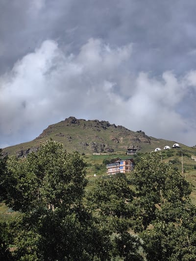 This image captures a mountainous landscape with several structures visible on the slopes. In the foreground, dense green trees dominate the lower part of the frame. Behind the trees, a steep, rocky mountain rises, covered in patches of green vegetation. Several buildings are situated on the mountain's side. One prominent building is a multi-story structure with a light-colored facade and blue-tinted windows. Further up the slope, there are several dome-shaped structures, possibly accommodations, and another smaller building. The sky above is filled with dramatic, gray clouds, suggesting overcast or impending weather conditions. The location is identified as Yamling, India. The scene appears to be a rural or semi-rural setting in a mountainous region. There are no people visible in the image.