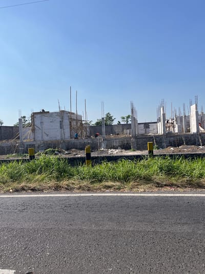 The image captures a construction site in the city of Purworejo, Indonesia, under a clear blue sky. The foreground is dominated by a paved road with a white line marking its edge, and beyond that, a strip of overgrown green grass. In the mid-ground, the construction site is visible, featuring the skeletal framework of a building. Numerous concrete pillars with rebar protruding upwards indicate the ongoing construction of multiple levels. White brick walls are being erected, and scaffolding made of bamboo poles and wooden planks supports the structure. Several workers are present at the site, engaged in construction activities. Two yellow and black striped posts stand along the edge of the construction area, possibly serving as markers. In the background, a grey brick wall encloses the construction site, with some trees and vegetation visible beyond it. The overall scene depicts an active construction project in an outdoor, sunny environment.