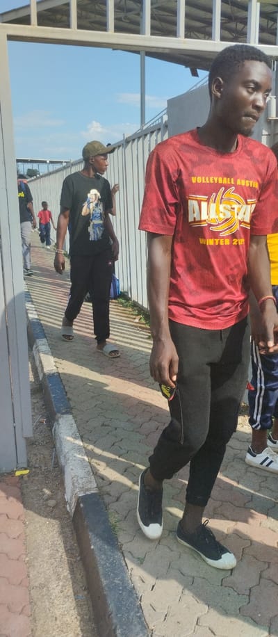 The image depicts a scene outside a sports facility in Kisumu, Kenya, specifically at the coordinates -0.066, 34.784. There are four people visible: a young man wearing a red shirt with the words "Volleyball Austin Allstar Winter 212" and black pants is the central figure, standing to the right of a man wearing a black shirt with a graphic print and black pants, who is walking away from the camera. A third man in a black shirt with the word "UN" visible is partially obscured by a metal gate on the left, and another man in a blue shirt is visible in the background. There is a black and white curb separating the paved walkway from a grassy area to the right. The walkway is made of interlocking brick pavers and has a slight slope downward. The metal gate appears to be an entryway to the sports facility, which is partially visible behind the people in the background.  The sky is blue and clear, with a few clouds. The overall tone of the scene is one of casual activity.