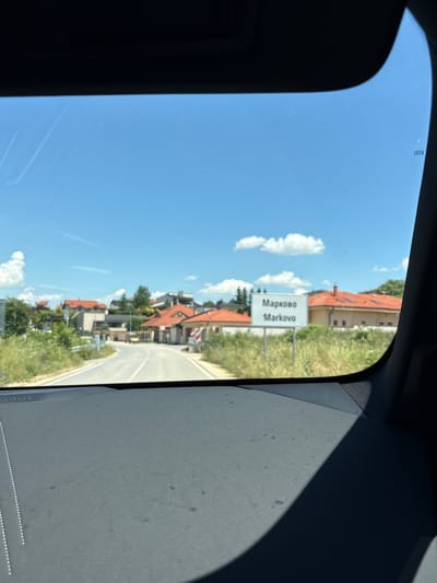 The image is a view through a car windshield of a road leading into the town of Markovo, Bulgaria. A signpost on the right side of the road indicates "Марково" and "Markovo." The landscape is rural, with houses with red tile roofs and green vegetation visible on either side of the road. The sky is clear blue with a few scattered clouds. The foreground of the image shows the dashboard of the car, with the word "AIRBAG" visible on the left.