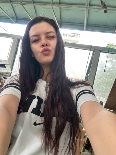 The image is a selfie taken by a young woman with long, dark hair and prominent eyelashes. She is wearing a white t-shirt with black stripes on the sleeves and a large black logo on the front. She is making a kissing face and winking.
The background suggests an outdoor or semi-outdoor setting, possibly a covered patio or balcony. There are metal beams and a green-painted ceiling structure above her. To the left, there are stacked cardboard boxes, one with text that appears to read "ANANAS". Through a window or open space to her right, trees and foliage are visible, indicating a green, possibly urban, environment. In the upper right corner of the image, a pigeon is perched on a wire.
The overall impression is a casual, candid shot taken in daylight. There is no specific information within the image that ties it to the city of Sliven, Bulgaria.
