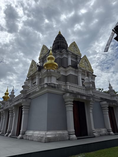 A low-angle, full shot captures the exterior of a temple in Chalong, Thailand, under a cloudy sky. The temple features a multi-tiered structure with gray walls, white decorative trim, and golden spires. Prominent architectural elements include large, dark wooden doors framed by thick, white columns. The upper sections of the temple are adorned with intricate gold-colored carvings and pointed roof structures. The surrounding area appears to be paved, with some grass visible in the lower right corner. The sky is filled with heavy, gray clouds, indicating overcast weather. There are no people visible in the image.