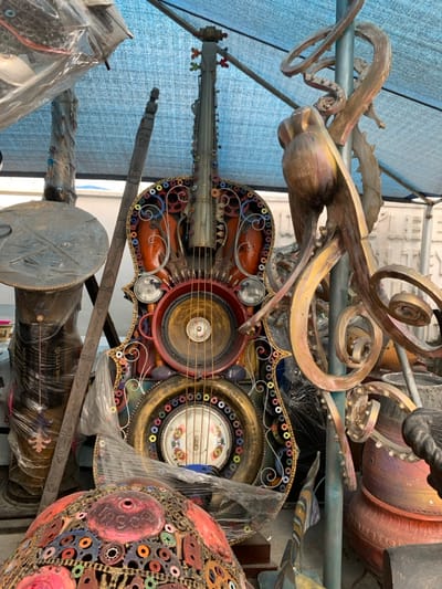 The scene depicts an outdoor art display or workshop area in Ikate, Nigeria. A large, approximately man-sized string instrument, resembling a cello or double bass, stands centrally, crafted from repurposed materials. Its body is primarily reddish-orange, intricately decorated with metallic gears, springs, bolts, and other industrial components, including two small headlights and a large speaker cone integrated into its soundboard area. The instrument's edges are adorned with numerous small, multi-colored rings. Below the speaker, a circular gold-colored frame surrounds a decorative white plate with patterns. To the immediate right, a large, multi-limbed sculpture, appearing metallic gold and bronze, resembles an octopus or abstract tendrils, affixed to a blue vertical support pole. Below this sculpture, several reddish-brown pottery items are visible. To the left, a tall, cylindrical object wrapped in clear plastic stands next to a dark, vertically oriented carved wooden staff. In the foreground, bottom left, a dome-shaped sculpture is partially visible, covered in a mosaic of small, colorful metallic rings and red painted sections, also partially wrapped in clear plastic. A light blue tarp provides overhead cover for the area. No individuals are present in the image.