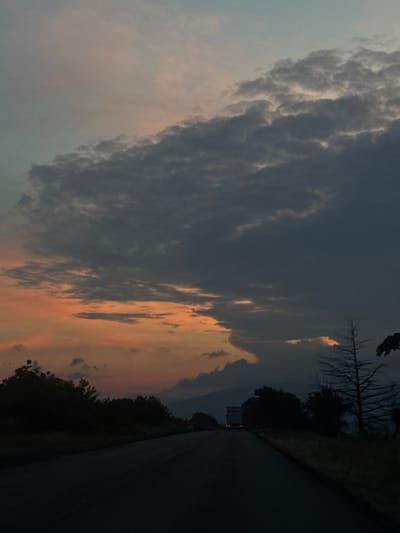 The image captures a dramatic twilight sky over a rural landscape, likely viewed from a moving vehicle. The sky is the dominant feature, characterized by a vast, heavy mass of dark grey clouds, some exhibiting distinct bulbous formations, occupying the upper left and central portions of the frame. Below and to the right of these dense clouds, the sky transitions to lighter, scattered clouds illuminated by warm orange, pink, and peach hues, indicative of a sunset or sunrise. Patches of pale blue-grey sky are visible through the cloud breaks.

On the right side of the frame, a dark asphalt road stretches into the distance. It appears to be a two-lane road, though markings are indistinct in the low light. The roadside is lined with dark silhouettes of trees and dense vegetation, including a prominent bare tree with stark branches standing out against the sky. In the far distance along the road, the red taillights of a vehicle, possibly a truck, are faintly visible. The distant horizon beneath the illuminated clouds shows dark, indistinct outlines, suggesting hills or mountains typical of the Orizari region in Bulgaria.

The scene conveys the atmospheric beauty of dusk or dawn, with the warm glow contrasting against the dark clouds and shadowed landscape. The weather appears heavily overcast but with dynamic lighting. No text is visible in the image.