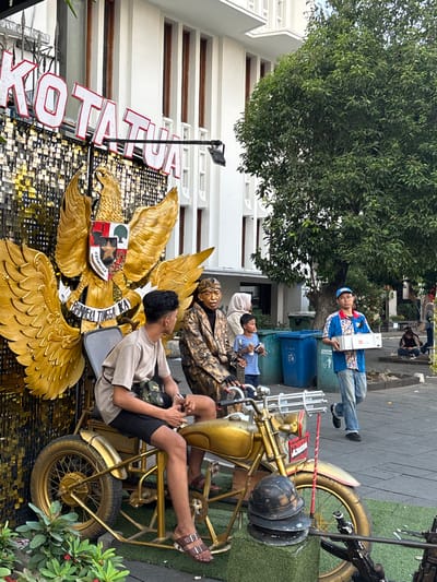 In the city of West Jakarta, Indonesia, a street scene unfolds with a prominent golden eagle emblem as the focal point. Two men, one young and casually dressed in a t-shirt and shorts, and an older man in a patterned jacket and headwear, are seated on a golden motorcycle. The younger man is on the left, facing to the right, while the older man is on the right, looking forward. Behind them, a large golden eagle statue with outstretched wings is mounted on a sequined backdrop that spells out "KOTA TUA" in large red letters. To the right of the motorcycle, a man in a blue shirt and cap walks carrying two white boxes. Several blue and green trash bins are visible in the background, along with a large green tree. The ground is paved with grey cobblestones, and some greenery is present in the foreground.
