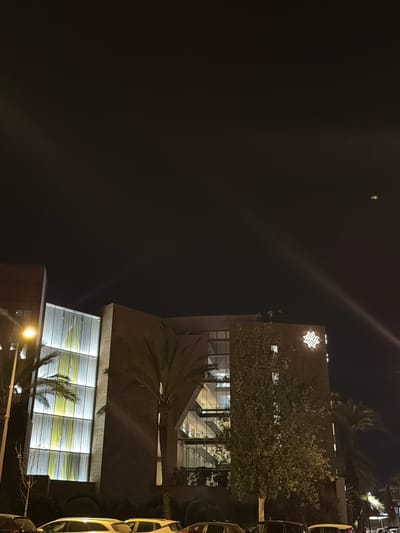 At night in Ibiza, Spain, a modern building with a glass facade is illuminated, revealing vertical yellow and white panels. The building, made of dark brown material, features a large, illuminated white star-shaped logo on its upper right side. Palm trees and deciduous trees are visible in front of and around the building, with streetlights providing additional illumination. Several cars are parked on the street in front of the building, and a few people are on the roof of the building, one of whom appears to be holding something. The sky above is dark and clear.