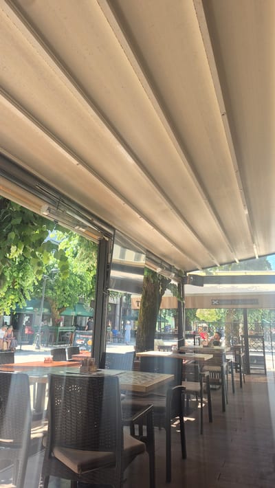 The image shows an outdoor seating area of a cafe or restaurant with a retractable awning providing shade. The awning is a light beige color and is extended, with visible support structures and heating elements attached underneath. The area is furnished with dark wicker chairs and tables with what appear to be patterned tabletops. Through the open sides of the covered area, a street scene is visible. Trees with lush green foliage line the street, and other businesses, some with green awnings, are present. Several people are seen seated at tables or walking on the street. In the background, there is a sign with the text "Scottish Pub Academia". The overall impression is a sunny day in an urban environment, likely a pedestrian area, with a relaxed atmosphere. Based on the signs and architectural style, the location is identified as the city of Cetinje, Montenegro.