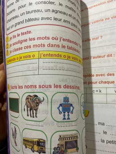 The image shows a page from a French workbook. The text includes sentences with words like "chameau," "taureau," and "agneau." Instructions prompt students to identify words where they hear certain sounds and categorize them in a table. The workbook also includes illustrations of a robot, bull, blackboard, bird, school bus, and a restaurant, asking students to write the names of each object below the images. Additional text is at the top of the page, in a barely visible form.