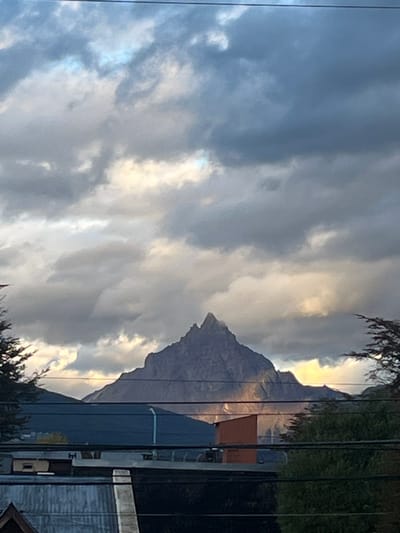 The image displays a static outdoor scene in Ushuaia, Argentina, centered on a prominent, dark grey mountain peak identifiable as Monte Olivia. The upper two-thirds of the frame are dominated by a partly cloudy sky, featuring dense, dark grey clouds intermixed with illuminated patches of pale yellow and bright white where sunlight penetrates. A single, thin, dark utility wire crosses horizontally at the extreme top. Monte Olivia rises sharply in the midground, its rocky, jagged form exhibiting areas of shadow and a distinct shaft of yellowish sunlight illuminating its lower right slope. Below the mountain, the foreground reveals urban architectural elements: a dark-colored building roof with a lighter ridge line, a small, rust-orange rectangular structure to its right, and a partial dark blue structure on the left. Multiple dark-colored utility wires extend horizontally across the lower third of the image, positioned above these foreground structures. Patches of dark green tree foliage are visible at the far left and right edges of the frame. No individuals or active subjects are present.