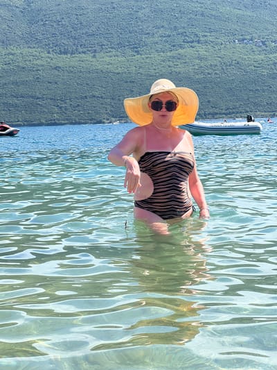 A woman stands in the clear, shallow water of the Bay of Kotor, near Baošići, Montenegro. She is wearing a tan and black tiger-striped one-piece swimsuit, a wide-brimmed straw hat, and sunglasses. Her left arm is extended slightly forward, with her hand open. In the background, a green, tree-covered mountainside rises from the water, with a few houses visible near the top. A white inflatable boat with "TENDER TO LANA" printed on its side is moored in the water, and a jet ski is visible to the left. The water is calm with ripples reflecting the sunlight.
