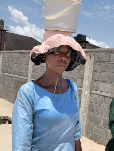 This image, taken in Alaguntan, Nigeria, presents an adult female positioned outdoors under a clear sky. She is depicted from the mid-torso up, wearing a light blue, long-sleeved dress featuring a subtle patterned texture. On her head, she balances a wide-brimmed, light pink sun hat with a black underside, over which a light-colored woven circular object is placed, topped by two stacked white plastic buckets. A light-colored chin strap from the hat hangs along her neck. She has small gold-colored hoop earrings and maintains a direct gaze with a neutral expression. A blue strap, possibly from a bag, is visible over her left shoulder. To the right of the frame, a segment of an individual wearing green clothing is partially visible. The background features a weathered concrete block wall. Above the wall, a dark-tiled roof is visible, along with two distinct elevated dark water tanks or containers supported by metal structures. The sky above is blue with scattered white clouds, and the ground in the foreground appears to be unpaved dirt.