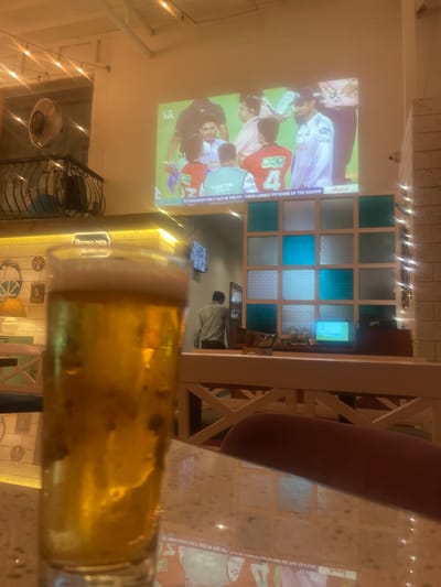 The scene is a restaurant in Mumbai, India, where cricket is being projected onto a wall, while a glass of beer sits on the table. The broadcast shows Gujarat Titans wearing jersey #4 and mentions "GT MANAGED ONLY 34/2 IN THE PP-THEIR LOWEST PP SCORE OF THE SEASON" and advertises brands like Amul, Astral Pipes and BKT tires. Other prominent text is "RETRO DEN". A fan and lights hang from the ceiling. Some people are partially visible in the background.