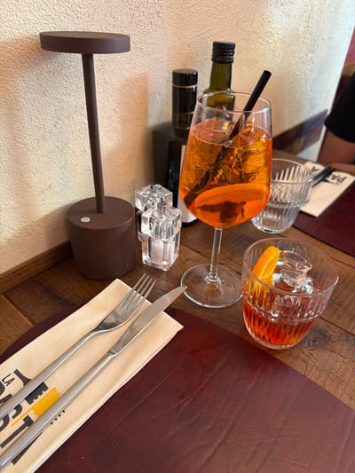 The image depicts a table at a restaurant in Como, Italy. On the table are two glasses filled with orange-colored beverages, likely Aperol spritzes, each garnished with an orange slice. One glass has a black straw. There are two small bottles of olive oil, a salt and pepper shaker set, and a set of silverware wrapped in a napkin. A brown, wall-mounted light fixture is visible. The table is made of wood, with a leather placemat under the silverware. The overall impression is of a dining experience in a restaurant setting.