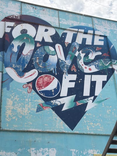 This is a vertical, eye-level medium shot of a faded, peeling outdoor mural on a light blue wall. The mural, centered and taking up most of the frame, features a large, stylized blue heart with the words "FOR THE LOVE OF IT" painted in white, blocky lettering. The heart contains abstract, flowing shapes in shades of blue, white, and red, with some sections resembling water splashes. The Pepsi logo, a red, white, and blue circle, is visible within the heart on the left side. The bottom portion of the mural features sharp, geometric lightning bolt-like shapes in dark blue, white, and teal. The wall itself is heavily weathered, with large areas of paint chipped away, revealing a lighter undercoat and textured concrete. The top of the wall has a metal frame and some overhead wires are visible against a cloudy sky. A wooden staircase is visible on the far right, ascending along the side of the wall. The overall impression is one of urban decay and a vintage advertisement. The mural is located on the side of a building in the city of Ulaanbaatar, Mongolia.