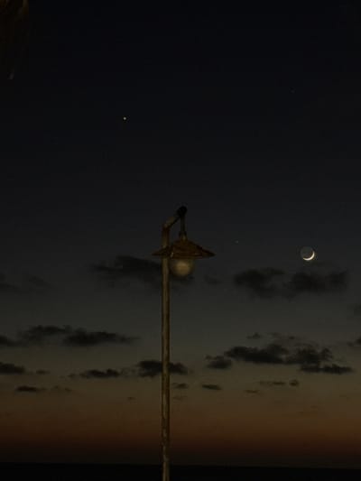 The image captures a dusky twilight sky over the city of Bat Yam, Israel. A solitary, weathered lamppost stands prominently in the foreground, its ornate metal fixture casting a warm glow from its bulb. In the distance, the faint silhouette of clouds drifts across the horizon, separating the darkening sky from the subtle gradient of orange and dark hues that mark the sunset. High in the sky, a sliver of a crescent moon is visible, accompanied by two faint pinpricks of light, likely stars or planets. The overall scene evokes a sense of quietude and the transition from day to night in a coastal urban setting.