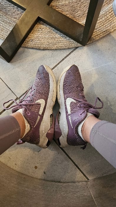 Two purple Nike sneakers with white swooshes are positioned heel-to-heel, one on top of the other, as if reflecting in a mirror. The shoes appear worn, with slightly soiled white soles and visible dirt. The person wearing the shoes is also sporting matching purple leggings and white socks. The backdrop consists of a gray stone floor with a rug and a dark brown table leg visible on the left side.