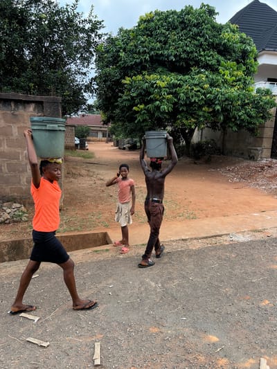 This image depicts a scene on a partially paved street or path in Nnobi, Nigeria, under an overcast sky. In the foreground, three individuals are present.
On the left, a female adolescent or young adult, positioned in profile, walks from left to right. She wears an orange short-sleeved top and a black skirt, with black flip-flops on her feet. Her hair is styled in braids. On her head, she balances two stacked, cylindrical green buckets.
In the center, slightly behind and to the right of the first person, a younger female child stands facing the viewer. She wears a light pink short-sleeved top and a beige pleated skirt, with pink flip-flops. She holds a dark-colored mobile phone to her right ear, appearing to be engaged in a call.
On the right, further back and behind the central child, an adolescent or young adult male walks from left to right, his back mostly to the camera. He is shirtless, wearing brown trousers with a dark belt, and dark sandals. He balances a single grey cylindrical bucket on his head, resting on a red and white patterned cloth.
The ground in the immediate foreground is a dark, gravelly asphalt mix with scattered light-colored debris. A concrete drainage ditch or gutter separates this paved section from a reddish-brown dirt path that extends into the background.
The background features dense green foliage from large trees on both sides, with several residential structures visible between them. On the far left, a light-colored block wall runs parallel to the path. In the distant center-left, a light-colored building with a reddish-brown corrugated roof and a white car is visible. On the far right, a larger, multi-story white building with a dark, possibly tiled, roof is partially obscured by trees.