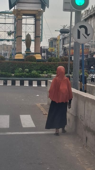 A person wearing an orange hijab and a long black skirt walks away from the camera, crossing a pedestrian crossing on an overcast day in Bireuen, Indonesia. The person is walking on the right side of the road, and a concrete barrier runs along the right side of the sidewalk. In the background, there is a tall monument with columns and a statue of a lotus bud at its center. Traffic lights and a traffic sign indicating a U-turn are visible, along with various buildings and a motorcycle in the distance. The scene appears to be a busy intersection in a city.