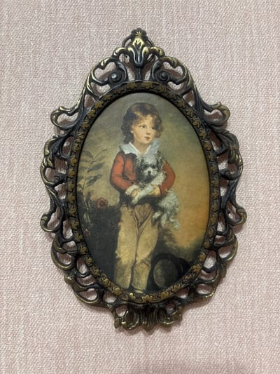 The media file shows an oval-shaped framed painting. The painting depicts a young boy dressed in historical clothing, possibly from the 18th century, wearing a reddish-brown jacket, a white collar, and breeches. He is holding a small, white, fluffy dog in his arms. The boy's expression is calm and gentle. He appears to be reclining or sitting in a naturalistic setting, with hints of foliage and a dark object, possibly a hat, in the background. The lighting suggests an indoor scene with soft, diffused light. The frame is ornate, made of metal with a dark finish and gold accents, featuring decorative scrollwork and leaf motifs. The background of the image is a light pink textured surface. There is no visible text in the image. The subject matter and style of the painting suggest a portrait from a historical period.