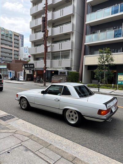 This detailed image captures an urban street scene in Daikoku, Naniwa Ward, Osaka, Japan, on an overcast day. The focal point is a white Mercedes-Benz 560 SL (R107 generation) hardtop coupe, equipped with aftermarket multi-spoke wheels, a black rear spoiler, and dual exhaust pipes, traveling eastbound on an asphalt road. A partial view of a driver is visible inside the vehicle. The rear license plate displays "560 SL" and the number "66".

To the left of the road, a concrete sidewalk features a square metal drain grate. Further left, a series of multi-story buildings line the street. In the middle ground, a low-rise commercial building with a brown facade houses a restaurant marked with Japanese characters for "鉄板焼" (Teppanyaki) and "お好み焼き" (Okonomiyaki). Outside this establishment, several bicycles are parked, alongside two white vending machines and two blue utility booths. In the background on the left, an apartment building with light-colored balconies and a distant building with glass-fronted sections are visible. A sign on a taller building in the distance partially reads "HOTEL RELIEF," and a blue sign with a stylized 'P' is also present.

On the right side of the image, a light-colored concrete sidewalk runs parallel to the street. A reddish-brown utility pole, equipped with cylindrical attachments and wires, stands prominently. Behind the pole, a tall, multi-story building with a grey facade and white grid-patterned balconies extends into the sky. Adjacent to this, a more modern, darker grey building features contemporary glass balconies, some appearing frosted or tinted blue. A small, verdant tree is planted in front of the modern building, and a green information board with Japanese text, partially mentioning "J-Leap," stands on the sidewalk.