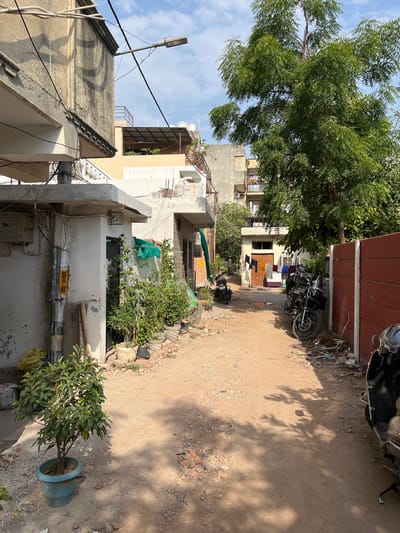 This is a vertical, eye-level shot of a narrow, dusty alleyway in Delhi, India. The scene depicts a residential area with multi-story buildings flanking the alley. The buildings are a mix of concrete and plaster, with some showing signs of age and wear. Laundry is hanging out to dry on lines strung between buildings, suggesting a domestic setting.

Several objects are visible, including motorcycles parked along the alley, and various pieces of debris and building materials scattered on the ground. A potted plant sits in the foreground on the right side of the image. A street lamp is visible on a pole extending from one of the buildings. The ground is composed of dirt, small stones, and some gravel.

The time of day appears to be daytime, with strong shadows cast by trees and possibly overhead structures, indicating sunlight. The weather seems to be clear and sunny, with a blue sky visible through the branches of a large tree on the left. The overall impression is one of a typical, somewhat cluttered, urban environment.

No people are visible in the image. There is some text on a yellow sign attached to a pipe on the side of a building, but it is too blurry to read. The scene is a common sight in many residential areas of Delhi, characterized by its narrow streets, closely packed buildings, and the visible signs of everyday life.