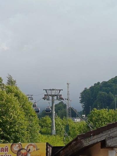 This image shows a ski lift with gondolas in motion on a bright, overcast day. The setting appears to be a mountainous, forested area, possibly a resort, given the ski lift. Lush green trees cover the slopes, suggesting a warmer season, likely summer or spring. The ski lift mechanism, with its tall support towers and cables, is a prominent feature. Several white gondolas are visible, suspended from the cables, indicating the lift is operational. In the foreground, a portion of a building with a weathered wooden roof and a yellow sign with Cyrillic text is visible. The sign displays images of coffee cups with latte art and a slice of citrus fruit, along with text that appears to be related to a cafe or beverage service. The overall atmosphere is serene and suggests an outdoor recreational environment.