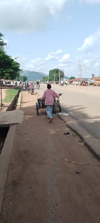 The image depicts a street scene in Gboko, Nigeria, likely in the late morning or early afternoon, given the bright sunlight and shadows. The weather appears to be clear with some scattered clouds. The foreground features a person from behind, pushing a two-wheeled cart. The person is wearing a pink long-sleeved shirt and light blue jeans. The cart appears to be made of metal and has some containers or items in it. The cart is on a dirt path next to a paved road.
The paved road is busy with traffic, including motorcycles and cars. Several other individuals are visible on the road and the sidewalk. On the left side of the road, there is a drainage channel and a row of trees. Further down the road, buildings and structures line the street, suggesting a commercial or residential area. In the background, a large, hilly landscape is visible under a bright, partly cloudy sky. Electrical poles and transmission towers are also present, indicating infrastructure. The overall scene conveys a sense of everyday life and activity in a bustling Nigerian town. There is no visible text in the image.