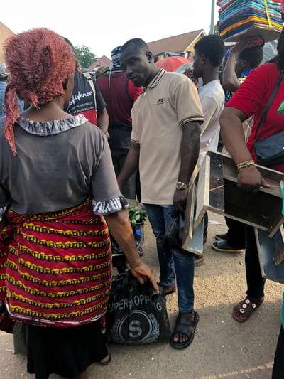 In a bustling outdoor market, likely in Nyanya, Nigeria, a group of people are engaged in commerce. The scene is vibrant and suggests a busy daytime setting, possibly a market day. The ground appears to be paved, characteristic of urban street markets.
The primary subjects are several individuals, including men and women. One person, wearing a red shirt, is partially visible on the left, with a stack of colorful textiles behind them. Another individual in a light-colored shirt is holding a wooden shelf unit with mirrored sections. A third person, wearing a black shirt with white lettering, is also present. A woman in the foreground, wearing a grey top and a vibrantly patterned red and yellow cloth, is looking down.
The activity appears to be the buying and selling of goods, with the shelf unit being a key object in focus, possibly for sale or being moved. The presence of textiles and the general market atmosphere indicate a commercial transaction.
Notable details include the gold watch worn by the person in the light-colored shirt, the beaded bracelet on the wrist of the person holding the shelf, and the "SUPERSHOP" text visible on a black bag. The weather seems to be bright, casting shadows, suggesting daylight. The overall impression is one of a lively and active marketplace.